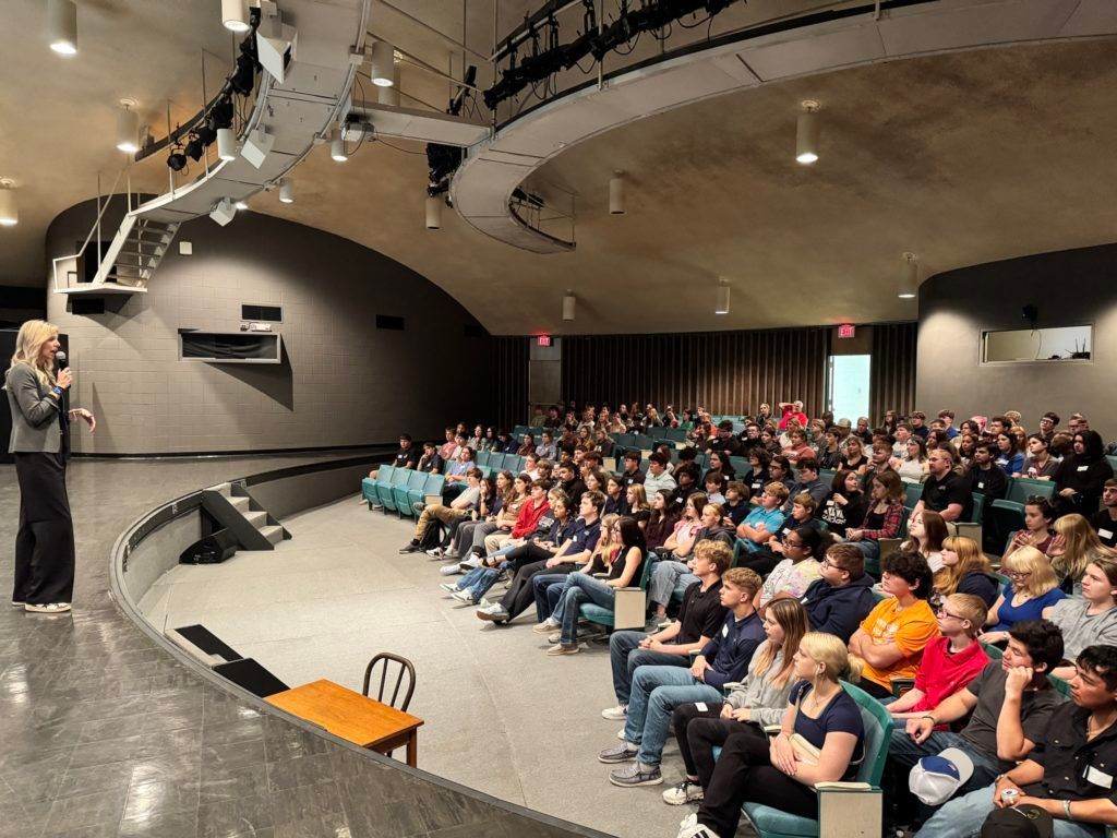 Speaker addressing a seated audience in an auditorium with curved ceiling and stage lighting