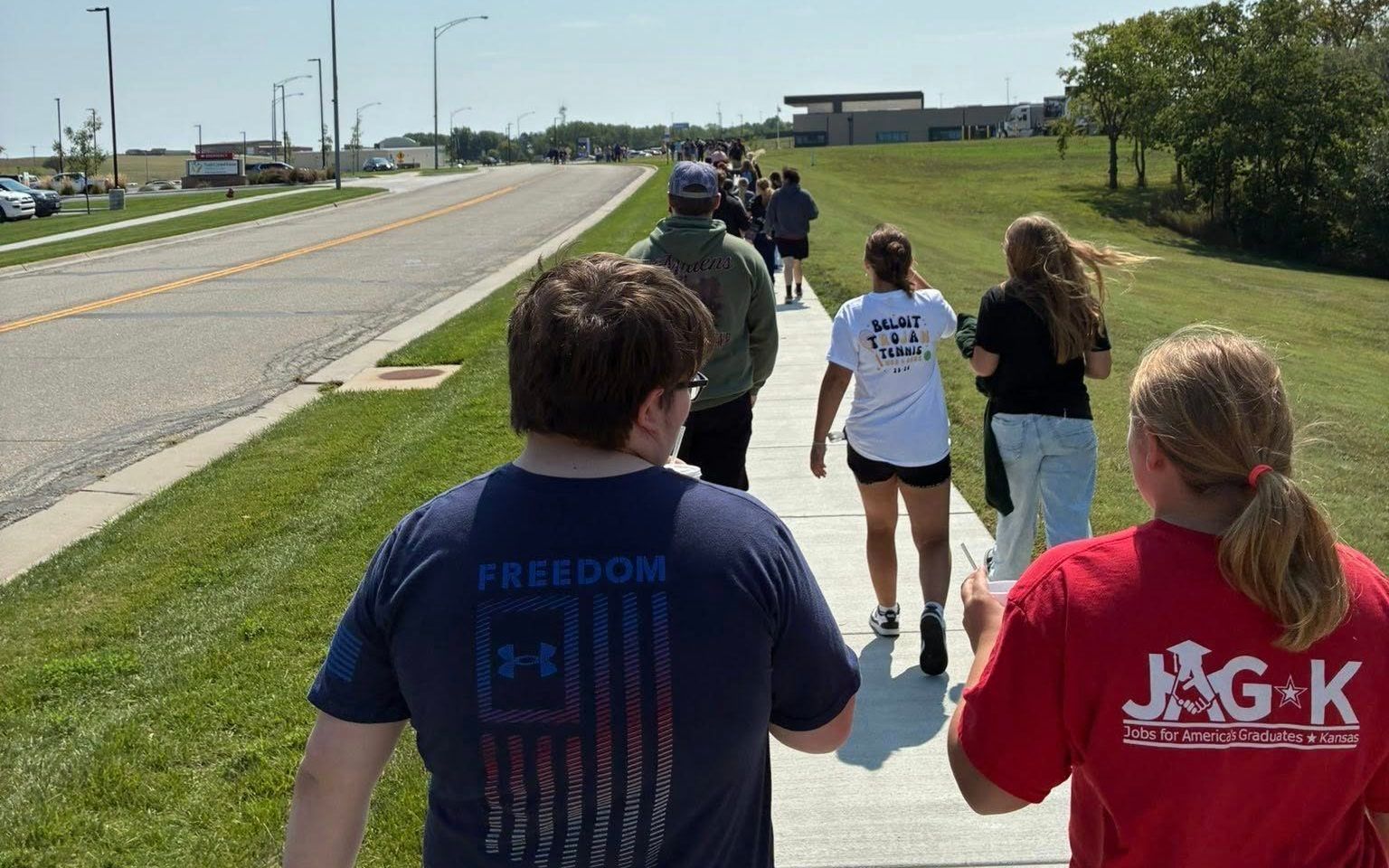 People walking along a roadside path on a sunny day, with cars on the road and open fields nearby.