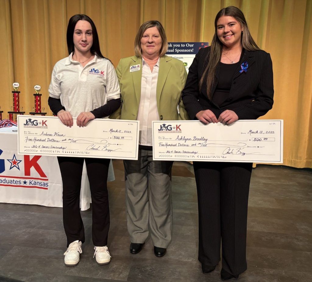 Three women holding oversized checks at an award ceremony, standing beside trophies and a table draped with a logo banner.