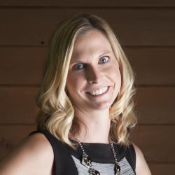 Smiling blonde woman in a black top and necklace against a wooden wall background