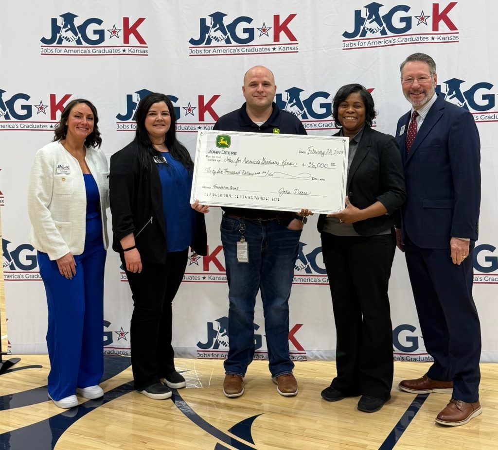 Five people at a J&G K event hold a large ceremonial check in front of a logo backdrop.