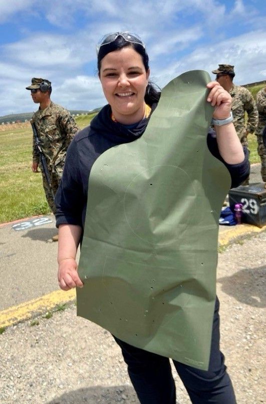 Smiling woman holding a large olive-green sheet outdoors with soldiers in camouflage behind her under a blue sky
