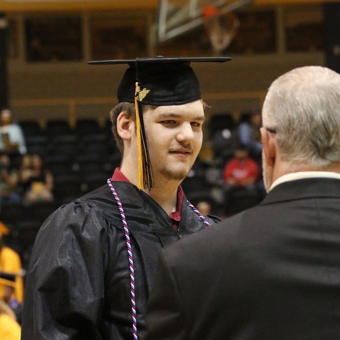 Graduate in black cap and gown with purple cords speaking to an older man at a ceremony