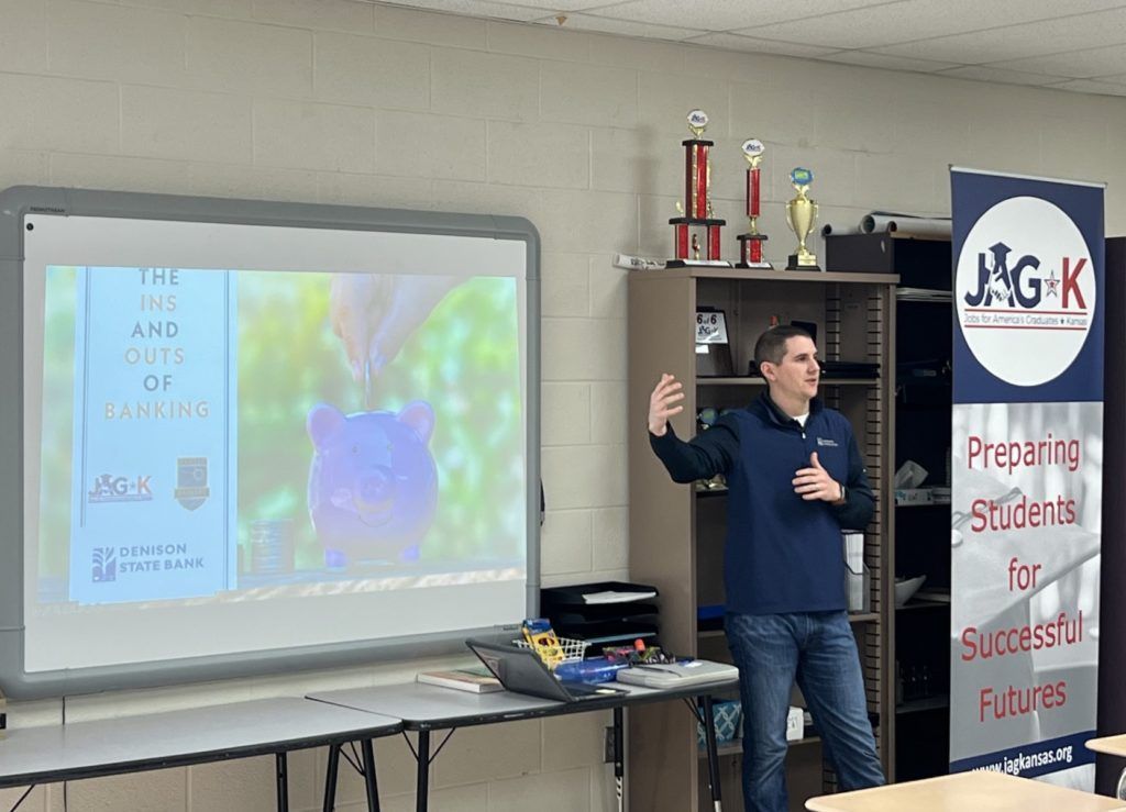 Presenter speaking beside a projector screen in a classroom, with a banner and trophies behind him.