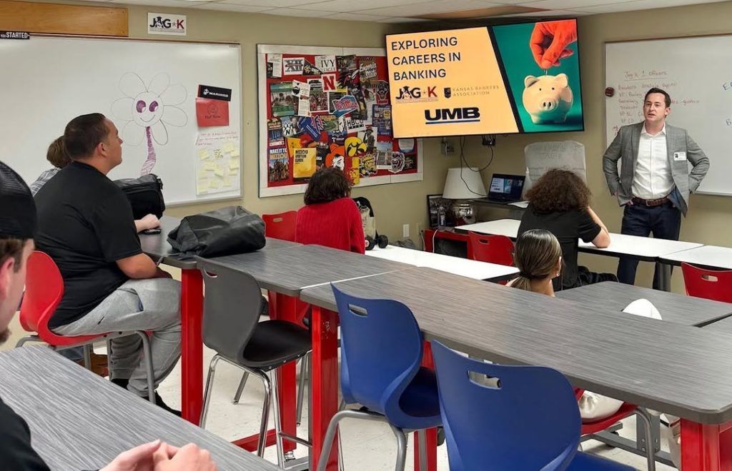 Classroom presentation with students seated at tables and a teacher speaking beside a screen.