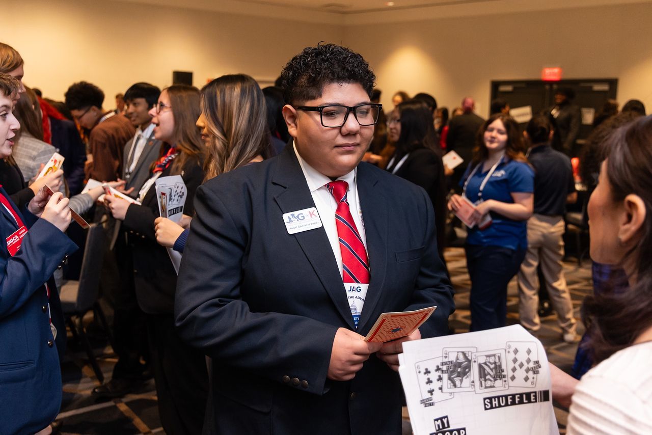 Man in a suit with a red tie holds cards at a crowded indoor event booth