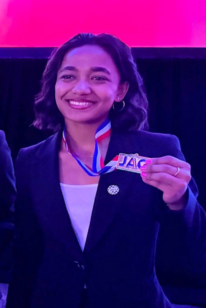 Smiling person in a dark blazer holding a medal with a red, white, and blue ribbon under purple lighting