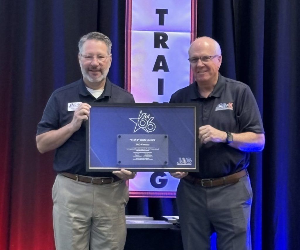 Two men hold a framed plaque onstage in front of a purple TRAI banner.