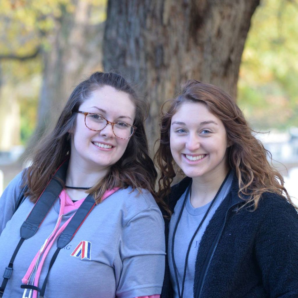 Two smiling women outdoors, standing by a tree in casual jackets and lanyards