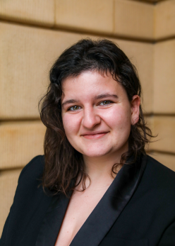 Portrait of a smiling person in a black blazer against a warm wood-paneled background