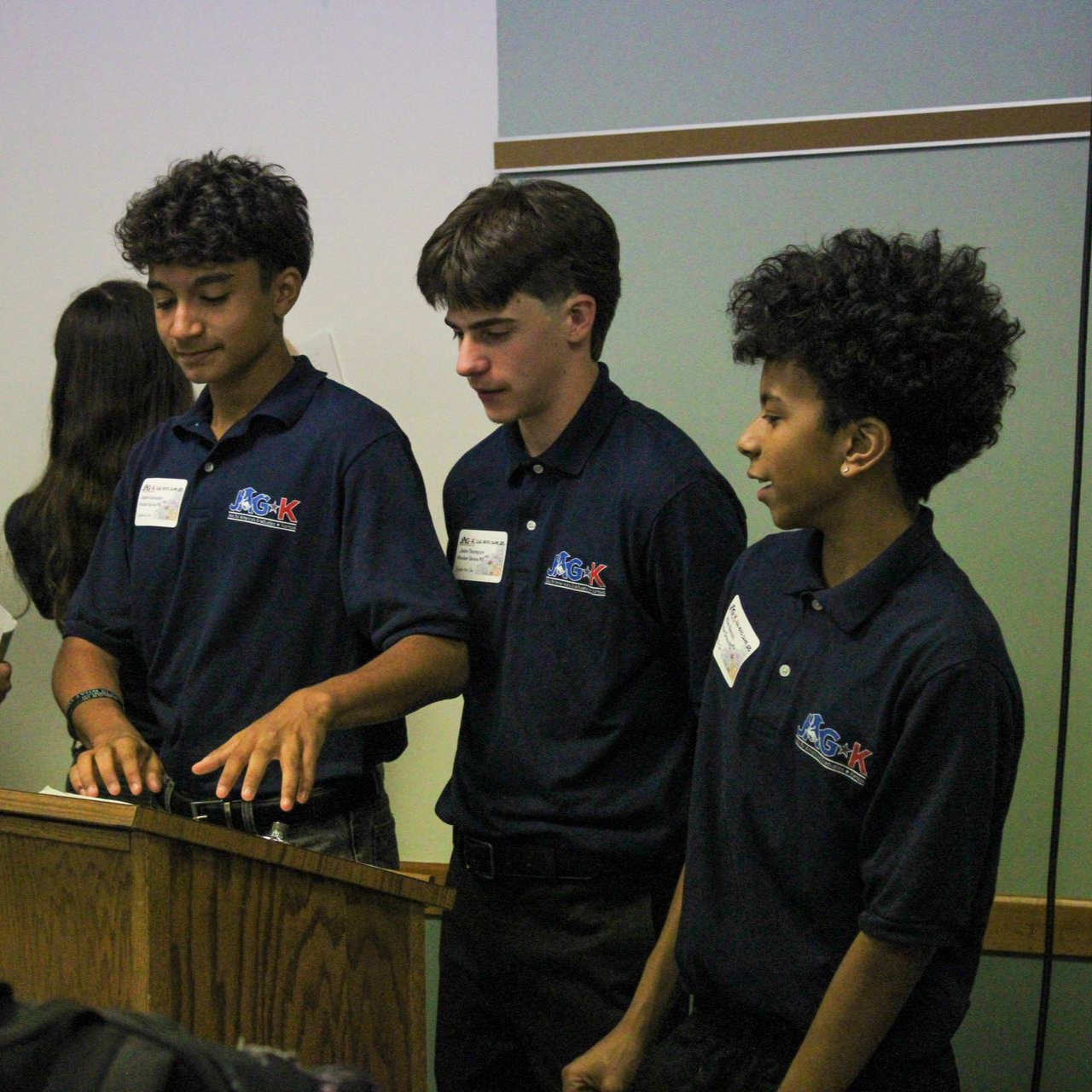Three students in navy shirts stand at a podium, one playing a keyboard during a classroom presentation.