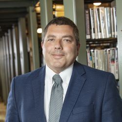 Man in a blue suit and tie standing indoors in front of bookshelves, facing the camera.