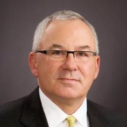 Studio headshot of a man in glasses, black suit, white shirt, and yellow tie on a gray background