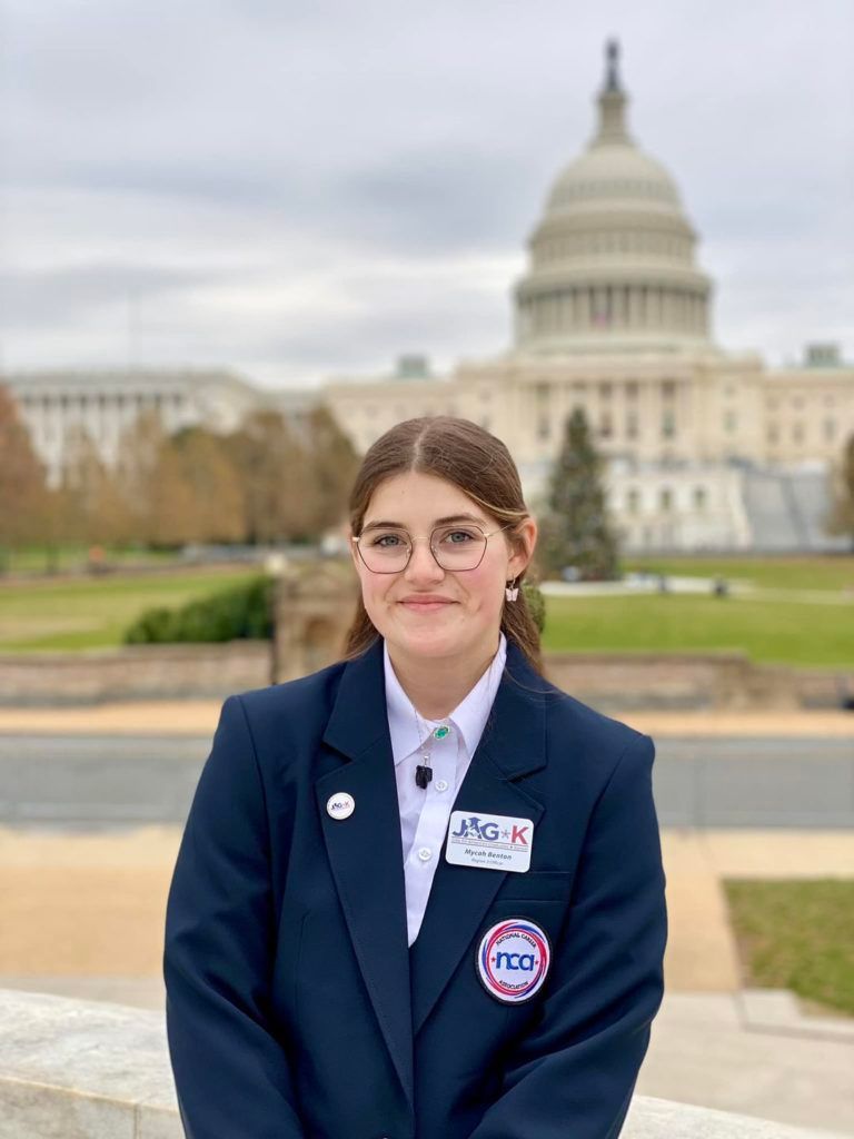Person in a navy blazer stands outdoors with the U.S. Capitol blurred in the background.