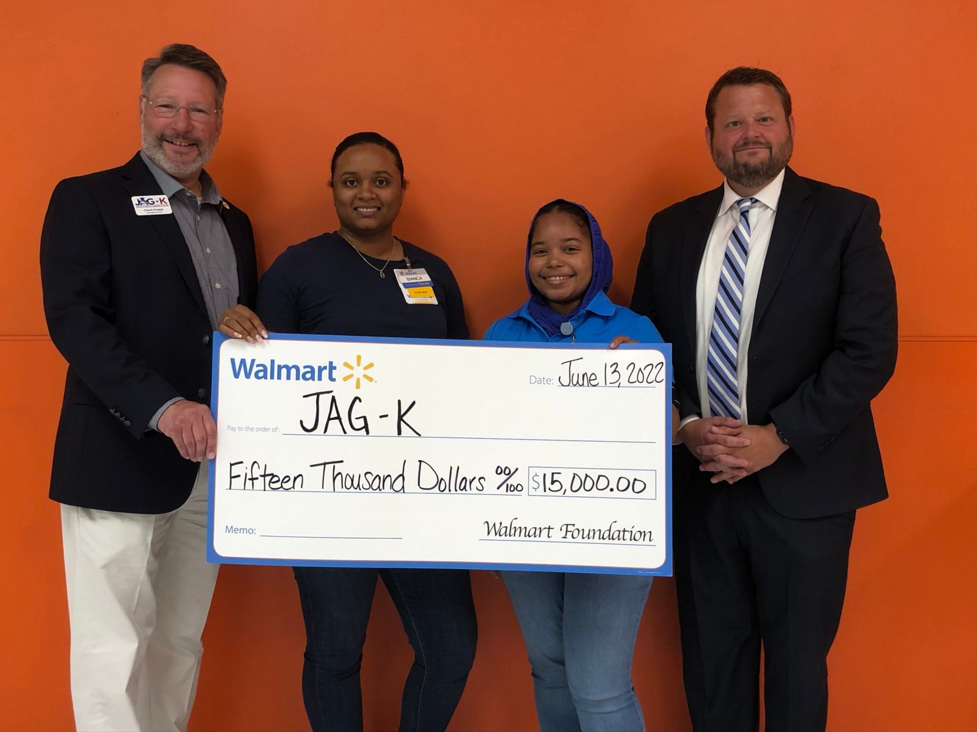 Four adults holding a large check in front of an orange wall, smiling for a photo.