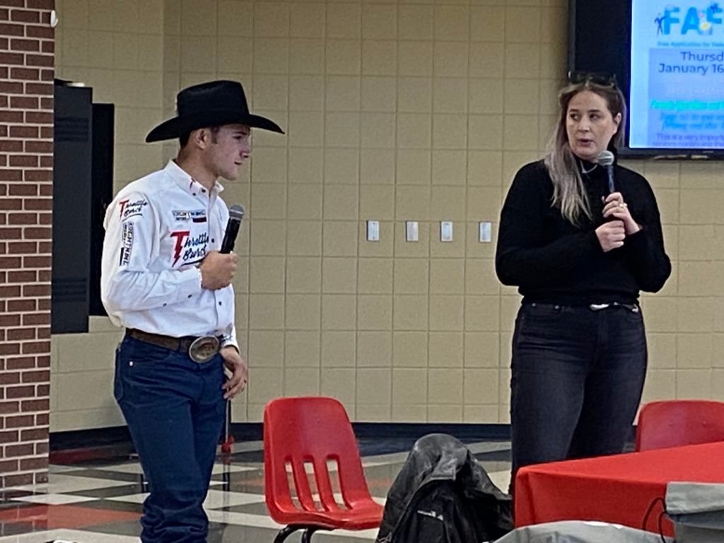Two people speaking indoors near red chairs, one holding a microphone and one wearing a cowboy hat.