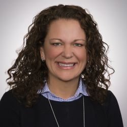 Smiling woman with curly dark hair in a black top and striped collar against a gray background