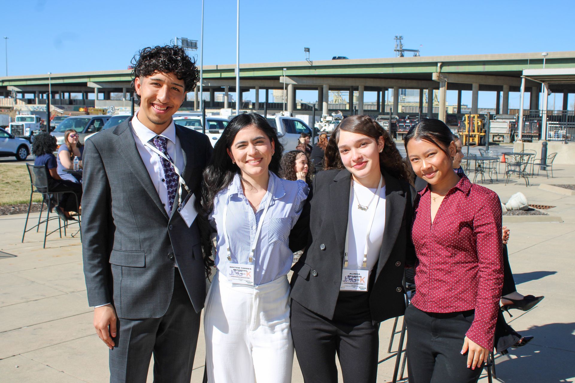Four people in business attire smiling outdoors in front of a transit station or parking structure.