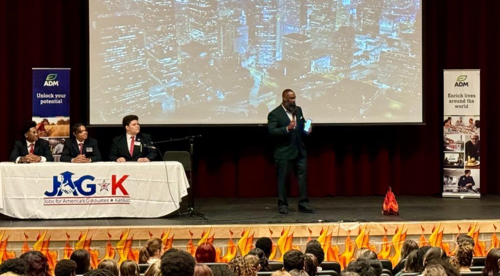 Stage event with speakers at a table and podium, large screen behind, audience seated in front.