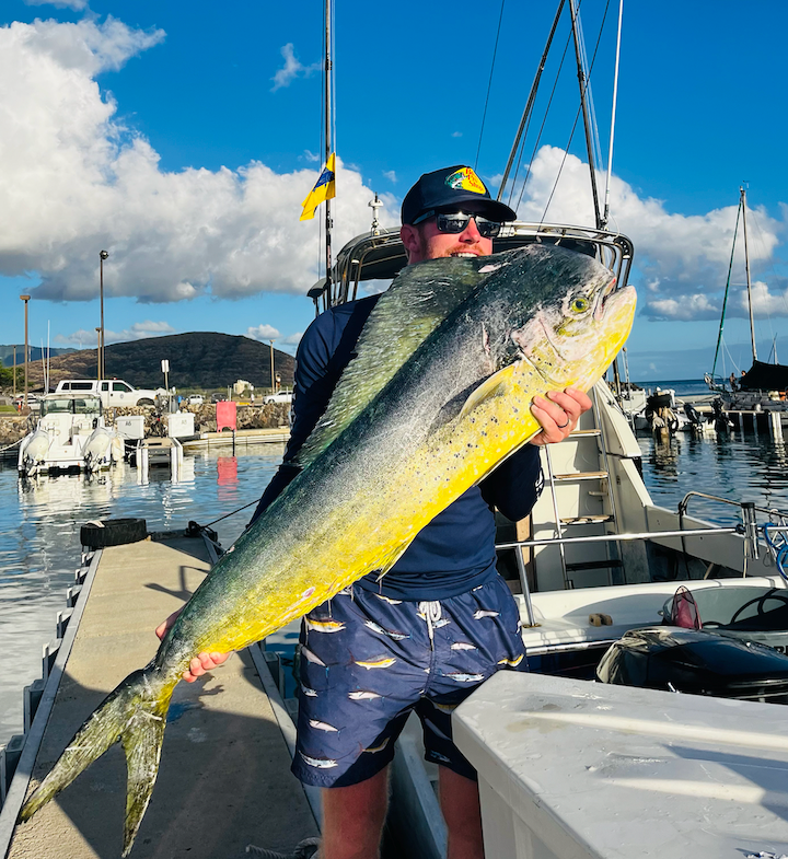 Person holding a large yellow-green fish on a docked boat in a sunny marina