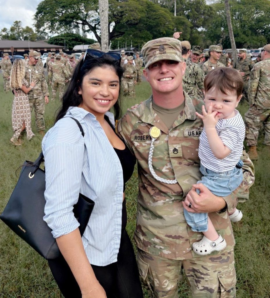 Smiling soldier in uniform holding a child, standing with a woman outdoors near a formation of troops