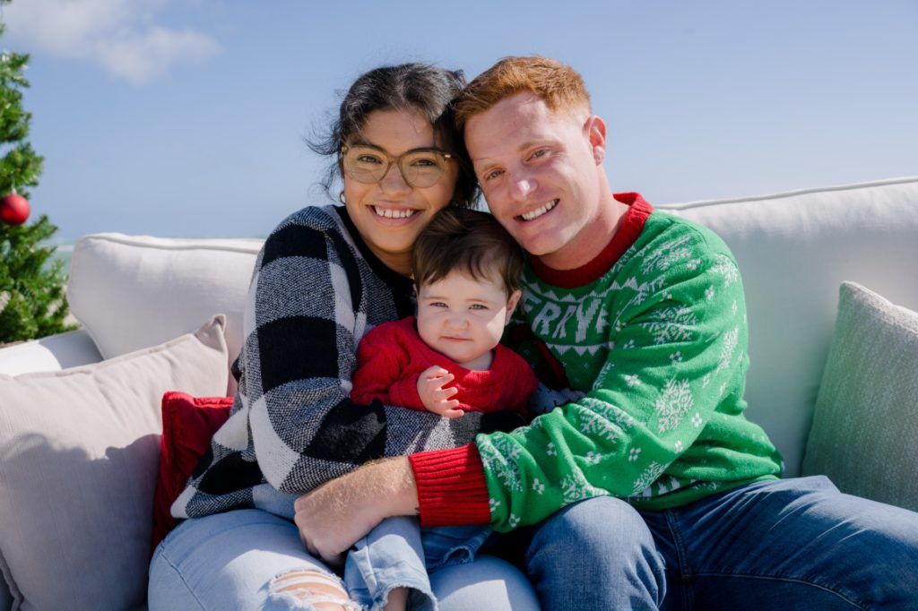 Family of three smiling on a couch outdoors, wearing cozy sweaters on a sunny day.
