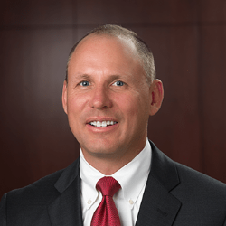 Professional headshot of a man in a dark suit and red tie against a dark background