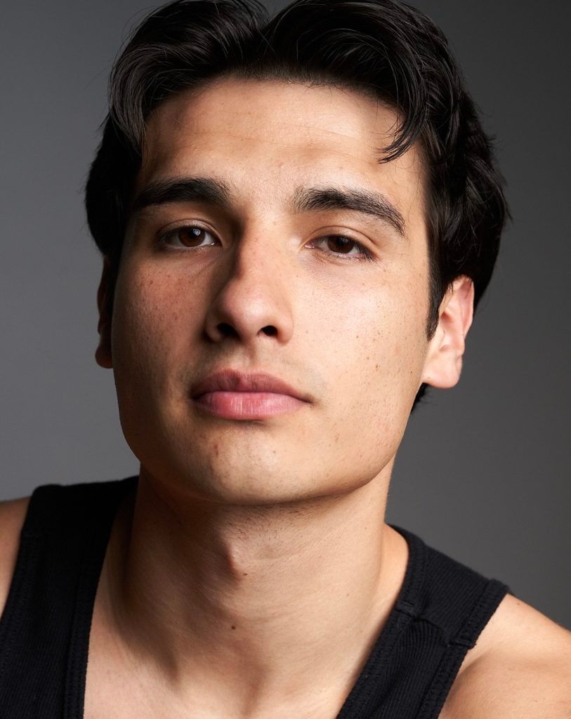 Close-up portrait of a person with dark hair wearing a black tank top against a gray background.