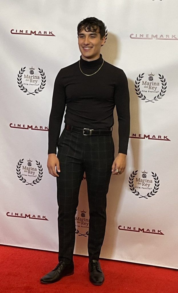 Person in black turtleneck and plaid pants posing on a red carpet with a branded backdrop.