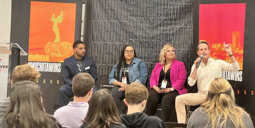 Panel discussion with five speakers seated onstage before an audience, flanked by colorful posters.