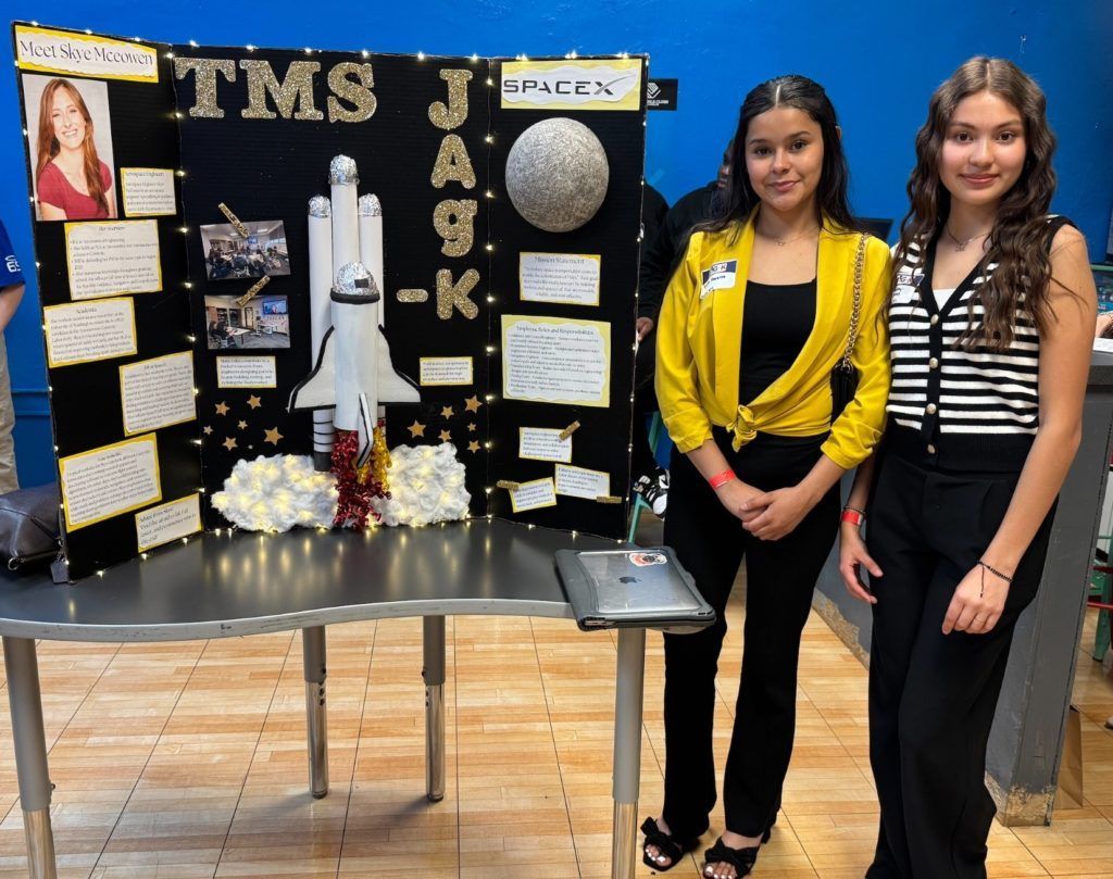 Two people standing beside a space-themed science fair display with a model rocket and planets.