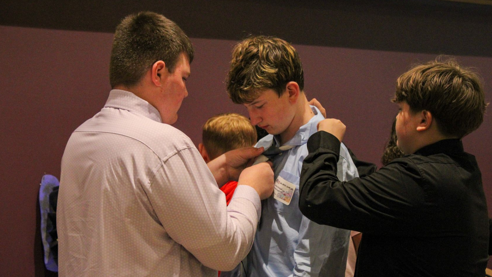 Three people helping adjust a young man’s tie or collar indoors, with one person holding a clipboard.