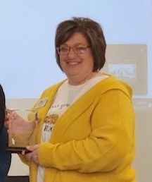 Woman in a yellow hoodie holding a trophy and smiling in front of a projector screen