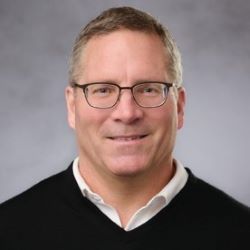 Portrait of a man wearing glasses and a black sweater over a white collared shirt, smiling against a gray background