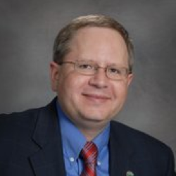 Studio headshot of a smiling man in glasses, wearing a navy suit, blue shirt, and red tie.