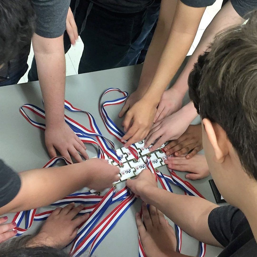 Hands of several people join over a table around a central object with red, white, and blue cords.