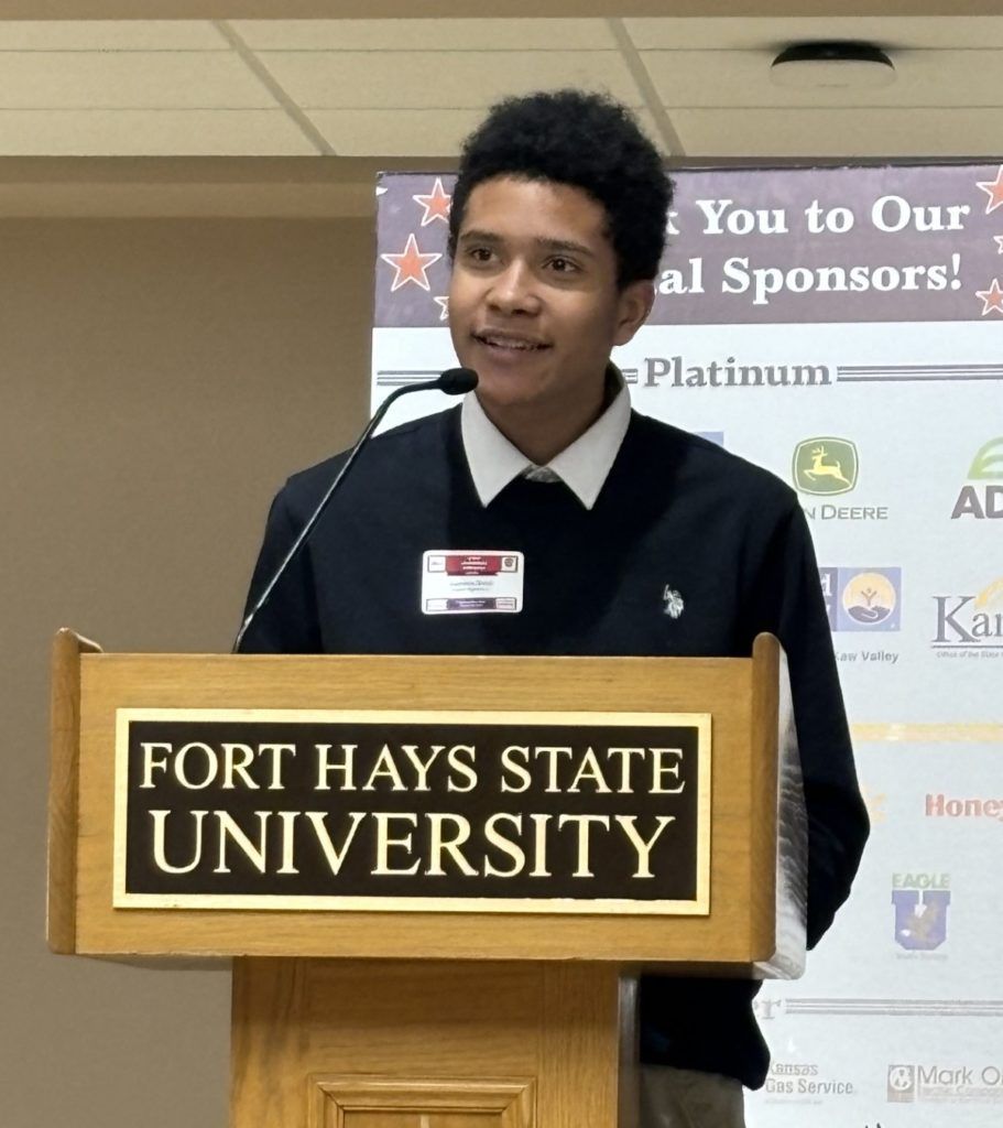 Speaker at Fort Hays State University podium during an indoor event, with sponsor banner behind.