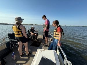 People in life jackets on a small boat, preparing to go out on calm water under a clear blue sky.