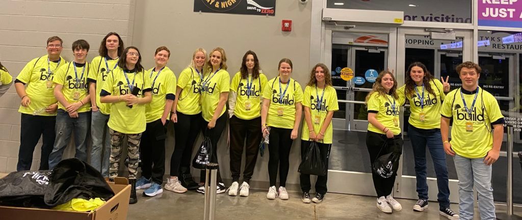 Group of people in neon yellow shirts standing indoors beside bags and a wall display