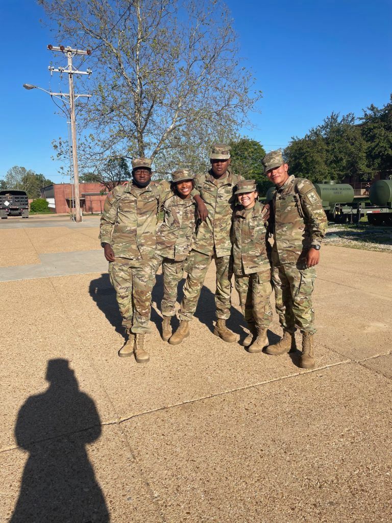 Four soldiers in camouflage uniforms posing together outdoors on a sunny day.