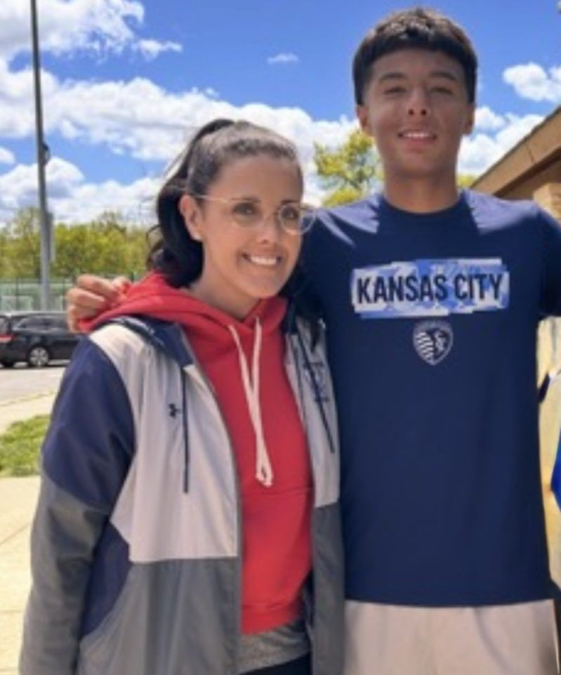 Two people smiling outdoors, one in a Kansas City shirt, under a bright blue sky.