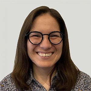 Smiling person with glasses and shoulder-length brown hair against a light gray background