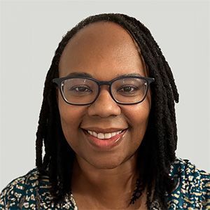 Smiling person with glasses and shoulder-length locs against a light background