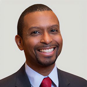 Smiling man in a dark suit, light blue shirt, and red tie against a plain background