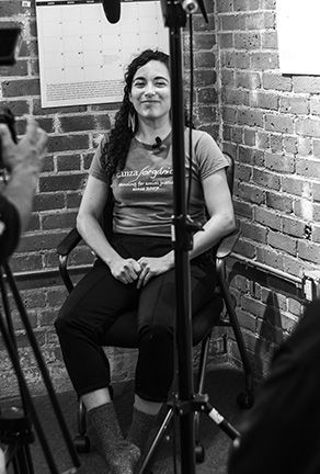 Woman seated indoors against a brick wall, smiling at the camera with a tripod in the foreground.