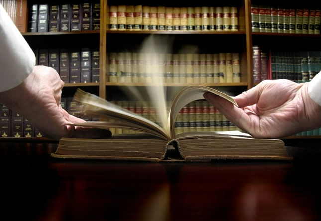 Two people are holding an open book in front of a bookshelf.