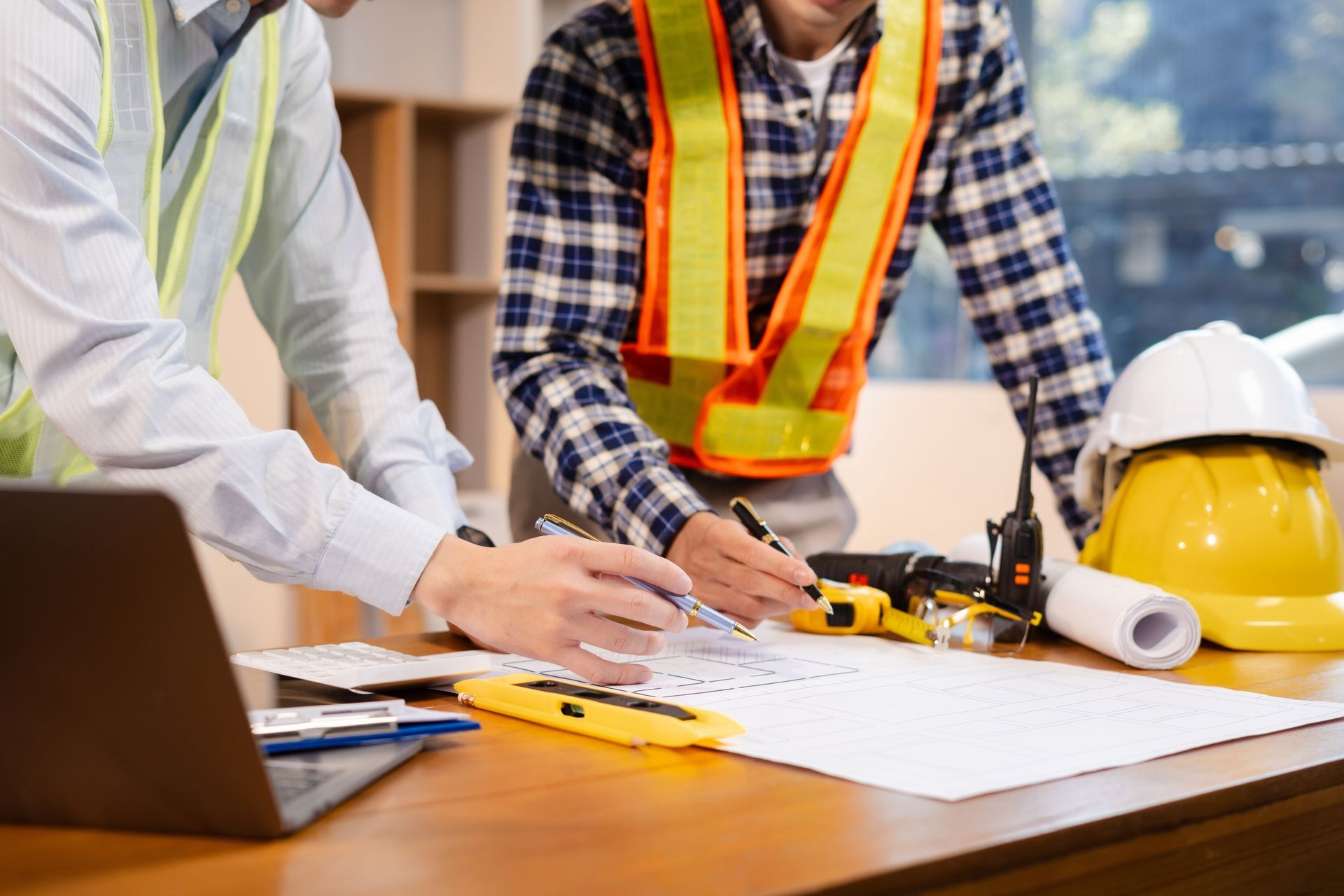 Two construction workers reviewing blueprints at a desk with tools and hard hats.