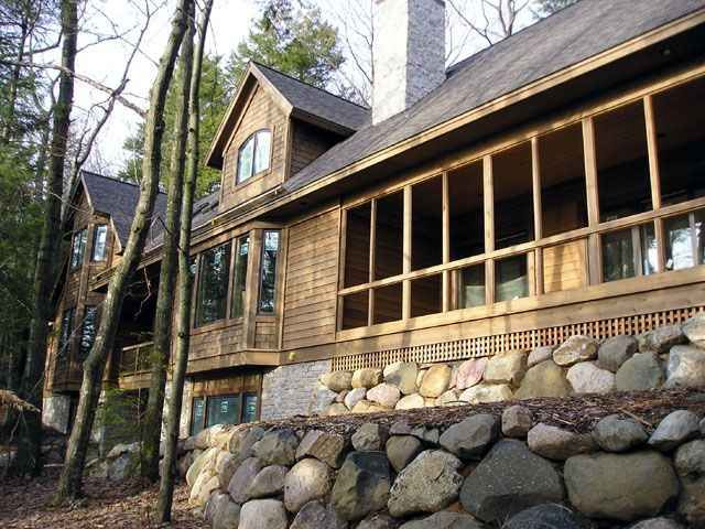 Wood-sided house with a screened porch and stone retaining wall; trees surround it.