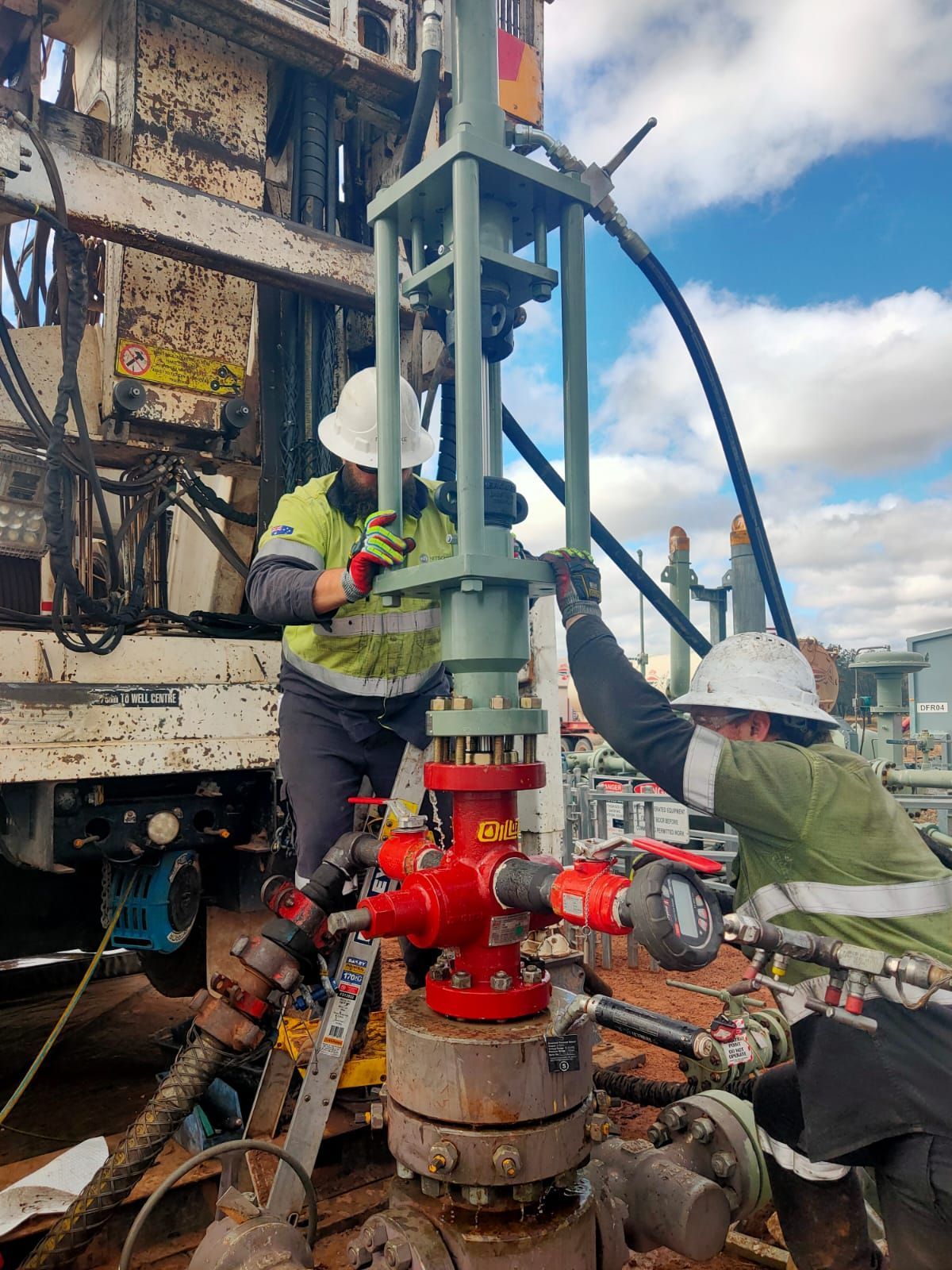 Workers in hard hats service a red industrial valve on drilling equipment outdoors.