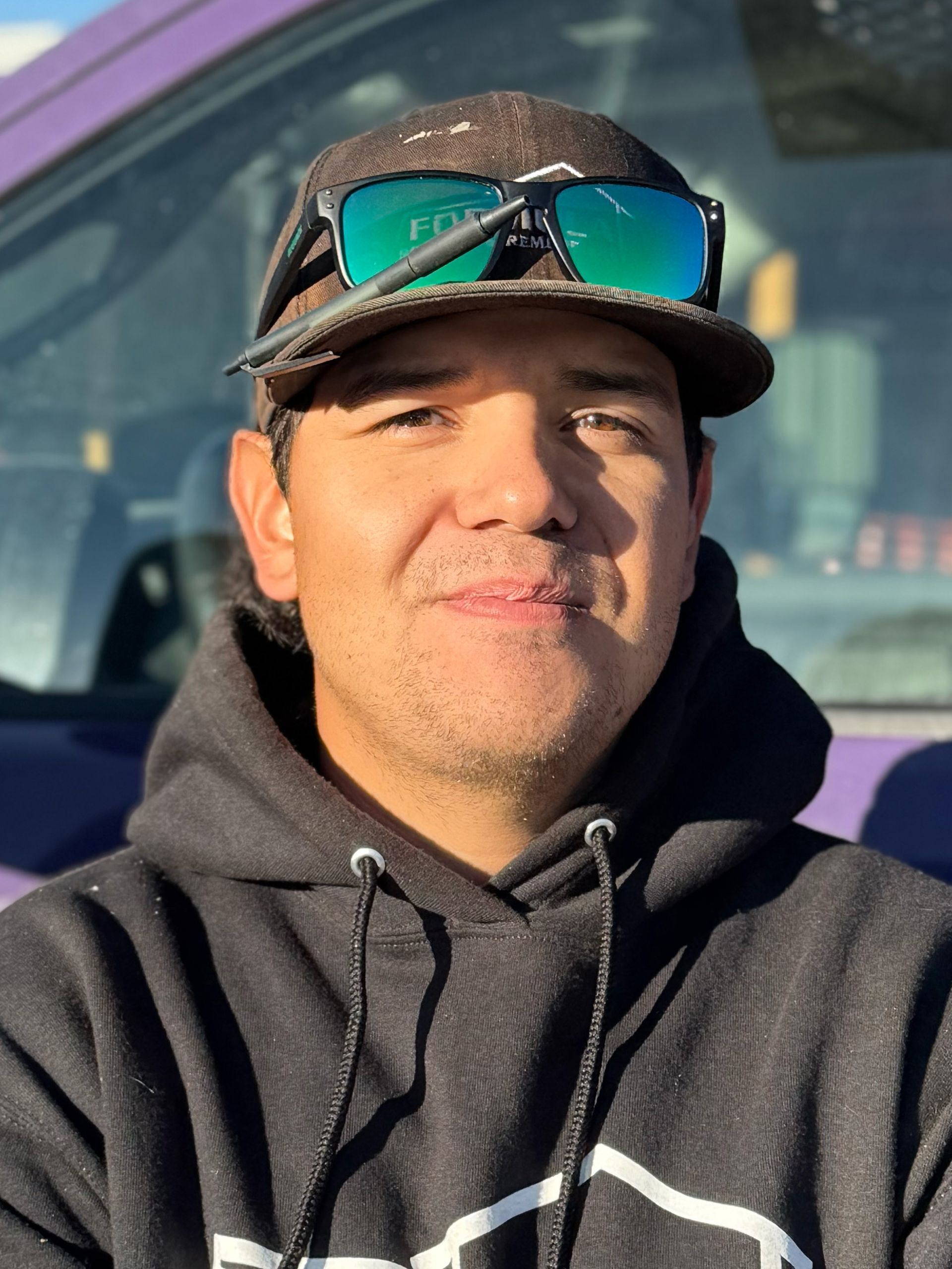 Man wearing a cap and sunglasses, in front of a purple vehicle. He has a neutral expression.